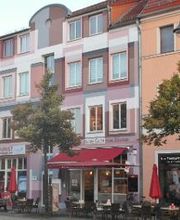 Rosa Gebäude, davor stehen Bäume. Unten im Gebäude ist ein Bäcker mit Sitzmöglichkeiten.