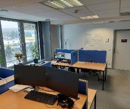 Raum mit mehreren Arbeitsplätzen mit jeweils 2 Bildschirmen, einem Bürostuhl, Tastatur und Maus. Links eine Fensterfront mit Zimmerpflanzen, im Hintergrund ein Kalender an der Wand. Der Boden ist mit blauem Teppich ausgelegt.