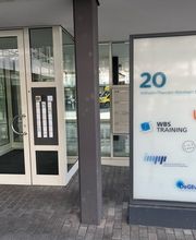 Außenansicht des Eingangsbereich vom Gebäude. Glas Eingangstür mit weißen Rahmen. Rechts steht eine Schildertafel der ansäßigen Unternehmen.