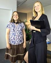 2 Frauen stehen neben einem Tisch. Frau rechts hält eine Klemmmappe. Im Hintergrund steht eine Zimmerpflanze und eine Stehlampe.