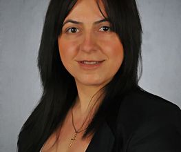 Eine Frau mit schwarzen Haaren, schwarzem Blazer und weißem Oberteil.