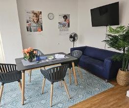Aufenthaltsraum mit Tisch, drei Stühlen und einer blauen Couch. Neben der Couch steht eine Zimmerpflanze.