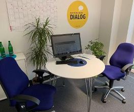 Ein kleiner Beratungsraum mit einem runden weißen Tisch, darauf ein Monitor, Tastatur und Maus. Zwei blaue Bürostühle stehen am Tisch. An der Wand ein gelber Kreis mit „Beratung heißt Dialog“, daneben ein Kalender und Pflanzen im Raum.