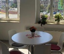 Tisch mit 5 Stühlen und einem Blumentopf. Dahinter 2 Fenster mit Pflanzen.