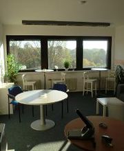 Pausenbereich mit3 Fenstern und Blick ins Grüne. Davor 2 Tische mit jeweils 2 Barhockern, einem größeren Tisch mit 2 Stühlen und einem grauen Sofa. An der Wand mehrere Pinnwände und Bilder. Ein Drucker steht im Raum