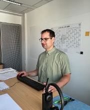 Ein Mann mit Brille und grünem Hemd sitzt an einem Schreibtisch. Vor ihm stehen ein Monitor, eine Tastatur und verschiedene Unterlagen. Im Vordergrund liegt ein Headset. An der Wand hinter ihm hängt ein Jahresplaner für 2024.