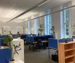 Schulungsraum mit mehreren Arbeitsbereichen, die durch blaue Trennwände voneinander abgegrenzt sind und mit PC-Bildschirmen, Tastatur, Maus und Headset ausgestattet sind. Fensterreihe mit Blick ins Grüne.