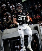 Ein American-Football-Spieler befindet sich in einer kraftvollen Sprungbewegung während eines Spiels. Emotion und Körperspannung stehen deutlich im Vordergrund.