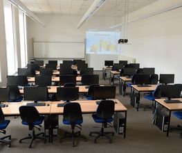Schulungsraum. Tischreihen mit Bildschirmen und blauen Stühlen. Die Wände sind rot und blau gestrichen. In dem Raum befindet sich ein Whiteboard und an der Wand ist eine Projektion vom Beamer zu sehen.
