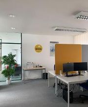 Ein Schulungsraum mit mehren Arbeitsplätzen bestehend aus 2 PC-Bildschirmen, Tastatur, Maus und Headset. Orang-graue Trennwände. Grüne Zimmerpflanze.