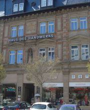 gelbes Haus mit dem Namen Haus des Handwerks. Im unteren Bereich des Hauses sind Schaufenster und davor stehen Autos. Im oberen Bereich sind mehrere Fenster und der Schriftzug Haus des Handwerks.