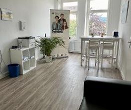 ein heller Aufenthaltsraum mit 2 großen Fenstern. Rechts steht ein weißer Tisch mit Bar-Hockern. Links steht ein Regal, auf dem ein Drucker steht. Daneben steht eine Zimmerpflanze und ein Roll Up der WBS GRUPPE. Im Vordergrund ist eine schwarze Couch