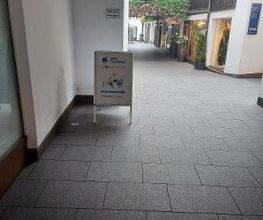 Schild mit Wegbeschreibung zum Eingang. Im Hintergrund mehrere Schaufenster.