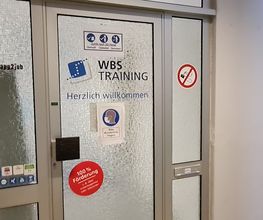 Glas-Eingangstür mit mehreren Aufklebern der WBS TRAINING wie einem Herzlich Willkommen Aufkleber und Hinweisen zur 100%igen Förderung.