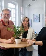 1 Mann und 2 Frauen stehen an einem Tisch mit Pflanze drauf. Alle 3 schauen freundlich in die Kamera. Neben ihnen steht eine weitere Pflanze.