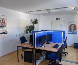 Raum mit 4 Arbeitsplätzen und 2 Türen. Arbeitsplätze mit jeweils 2 Bildschirmen, einem Bürostuhl, Tastatur und Maus. Links und rechts hängen Plakate der WBS GRUPPE und WBS TRAINING. 