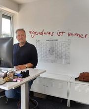 Ein Bild eines Mitarbeiters der an seinem höhenverstellbaren Schreibtisch steht und am Computer arbeitet. Im Hintergrund ist ein Schriftzug auf der Wand der sagt „irgendwas ist immer ...“