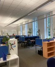 Schulungsraum mit mehreren Arbeitsbereichen, die durch blaue Trennwände voneinander abgegrenzt sind und mit PC-Bildschirmen, Tastatur, Maus und Headset ausgestattet sind. Fensterreihe mit Blick ins Grüne.