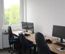 Arbeitsbereich ausgestattet mit 2 Bildschirmen, einer Tastatur, Maus und Headset. Fenster mit Blick ins Grüne.