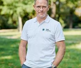 Stefan Schrader steht im weißen T-Shirt mit WBS GRUPPE Logo in der grünen Natur und lächelt in die Kamera.