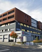 Außenansicht des Gebäudes. Im Erdgeschoss befinden sich mehrere Garagen und auf der grauen Fassade ist ein rotes Kreuz  zu sehen. Die darüberliegenden Etagen haben eine schwarze und rote Fassade sowie viele Fenster und Balkone.