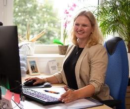 Eine Frau in beigem Blazer sitzt lächelnd an einem Schreibtisch mit Monitor, Tastatur und Unterlagen. Im Hintergrund stehen Pflanzen, eine Orchidee und ein Foto auf der Fensterbank. Durch das Fenster sind unscharf grüne Bäume zu sehen. Der Raum ist hell.