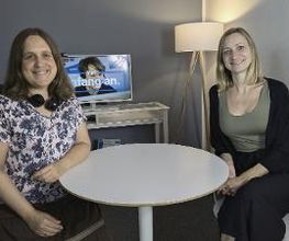 2 Frauen sitzen am Tisch und lächeln in die Kamera. Im Hintergrund ein Bildschirm auf einem Regal. Daneben eine Stehlampe.