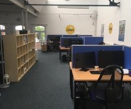 Schulungsraum mit mehreren Arbeitsplätzen mit jeweils 2 Bildschirmen, einem Bürostuhl, Tastatur, Maus und Headset. Links steht ein Holzregal.