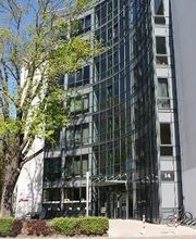 Außenansicht vom Gebäude. Helle Fassade mit vielen Fenstern. Im Vordergrund steht ein großer Baum.