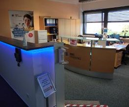 Empfangsbereich mit 2 Theken, einer mit LEDs und Händedesinfektion, die andere aus Holt mit Glasplatte. Hinter der Holztheke sind 2 Arbeitsplätze und mehrere Holzschränke