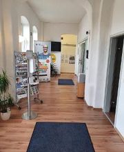 Flur zu verschiedenen Räumen. Links ein Flyerständer und dahinter ein Roll Up Banner. Rechts mehrere Türen und ein Schrank mit Drucker.