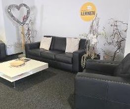 Wartebereich bestehend aus einem schwarzen Ledersofa und zwei Ledersessel mit einem Tisch in der Mitte. Im Hintergrund steht ein Herzaufsteller.