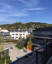 Sonnige Aussicht vom Balkon mit Blick auf einen Hof mit Grünflächen und Bäumen. Im Hintergrund ein weißes Nachbarhaus und Parkplätze hinter dem ein Berg mit Wald ragt.