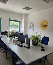Mehrere Arbeitsbereiche, die mit 2 PC-Bildschirmen, Tastatur, Maus und Headset ausgestattet sind. Einige grüne Topfpflanzen stehen auf den Tischen und im Hintergrund befindet sich ein gelbes Wandtattoo mit der Aufschrift „Fürs Leben lernen.“
