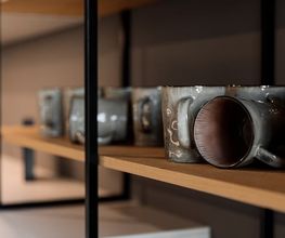 Detailaufnahme eines modernen, offenen Küchenregals aus hochwertigem Holz mit handgefertigten Keramiktassen in natürlichen Grau- und Brauntönen. Die Kombination aus dunklem Metallrahmen, warmem Holz und minimalistischer Keramik unterstreicht das elegante, zeitgemäße Küchendesign.