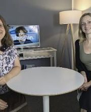 2 Frauen sitzen am Tisch und lächeln in die Kamera. Im Hintergrund ein Bildschirm auf einem Regal. Daneben eine Stehlampe.