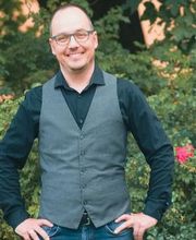 Mann mit Brille und Hemd steht draußen vor grünen Pflanzen und lächelt in die Kamera.