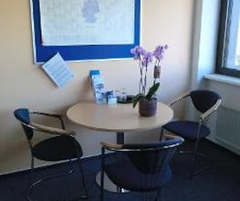 Bild von einer Sitzecke. 1 runder Tisch und 3 Stühle. Auf dem Tisch steht eine lavenderfarbene Topfpflanze und an der Wand hängt eine blaue Pinnwand an der ein Kalender befestigt ist.