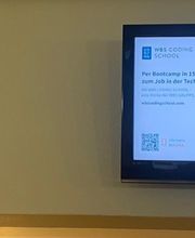Teilnehmerinforamtionen auf einem Fernseher, der an einer Wand im Eingangsbereich hängt. Daneben steht geschrieben WBS TRAINING Empfang mit Richtungspfeilen.