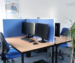 Mehrere Arbeitsplätze mit jeweils 2 Bildschirmen, einem Bürostuhl, Tastatur und Maus. An der Wand hängt ein WBS TRAINING Plakat.