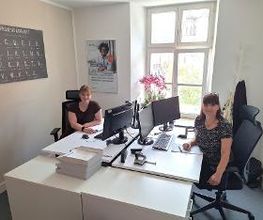 2 Frauen sitzen am Arbeitsplatz und schauen freundlich in die Kamera. Ihre Tische sind ausgestattet mit 2 Bildschirmen, Tastatur und Maus. Zwei Plakate hängen an der Wand.