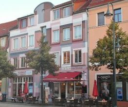 Rosa Gebäude, davor stehen Bäume. Unten im Gebäude ist ein Bäcker mit Sitzmöglichkeiten.