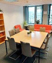 Pausenraum mit großem Tisch mit 8 grauen Stühlen. Links steht ein Holzregal mit Büchern und Deko, im Hintergrund ein rot/oranges Sofa und ein Sessel. Auf dem Fensterbrett stehen mehrere Zimmerpflanzen. 