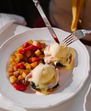 A close-up of hands using a knife and fork to cut into poached eggs Benedict topped with hollandaise, served alongside crispy diced roasted potatoes.