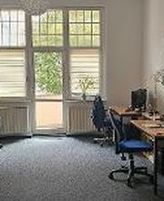 Schulungsraum mit links und rechts mehreren Arbeitsplätzen. Jeweils mit 2 Bildschirmen, einem Bürostuhl, Tastatur, Maus und Headset. Am Ende des Raums mehrere Fenster und eine Tür zum Balkon. An den Wänden mehrere Plakate.