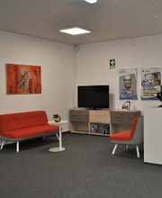 Empfangsbereich mit rotem Sofa und Sessel, daneben ein Regal mit TV. An den Wänden hängen Plakate und ein rotes abstraktes Gemälde.