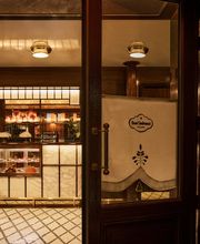 A wooden entrance door with a white branded curtain offers a view into a warm, warmly lit bakery and café with mosaic tile flooring.