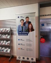 Flur mit Glastür zum Seminarraum. Links steht ein Flyerständer und ein Roll Up Banner der WBS GRUPPE. Rechts die Glastür mit Aufkleber "Herzlich Willkommen"