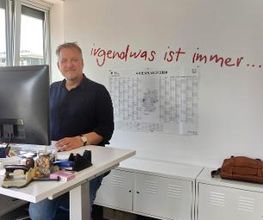 Ein Bild eines Mitarbeiters der an seinem höhenverstellbaren Schreibtisch steht und am Computer arbeitet. Im Hintergrund ist ein Schriftzug auf der Wand der sagt „irgendwas ist immer ...“