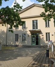 Außenansicht vom Gebäude. Helle Fassade mit weißen Fensterrahmen. Vor dem Gebäude ist eine Grünfläche mit Bäumen und einer Bank.